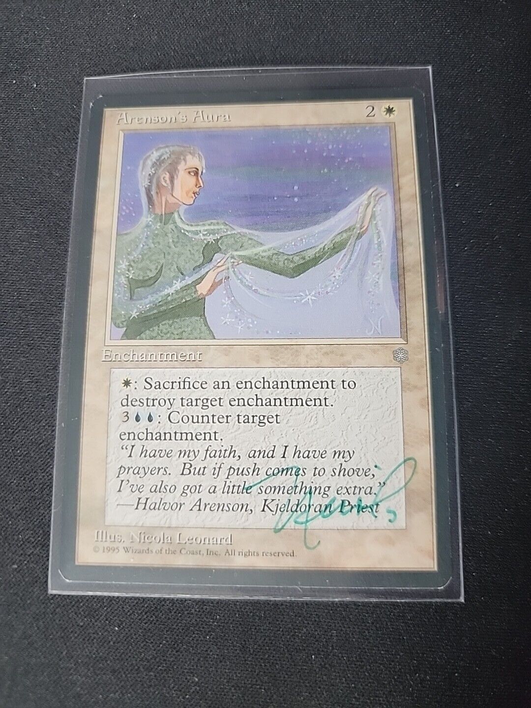 An image of a Wizards of the Coast Magic: The Gathering card named "Arenson's Aura," featuring artwork of a female figure holding a glowing star within a mystical backdrop, showing a signature and a heartfelt message on the card.